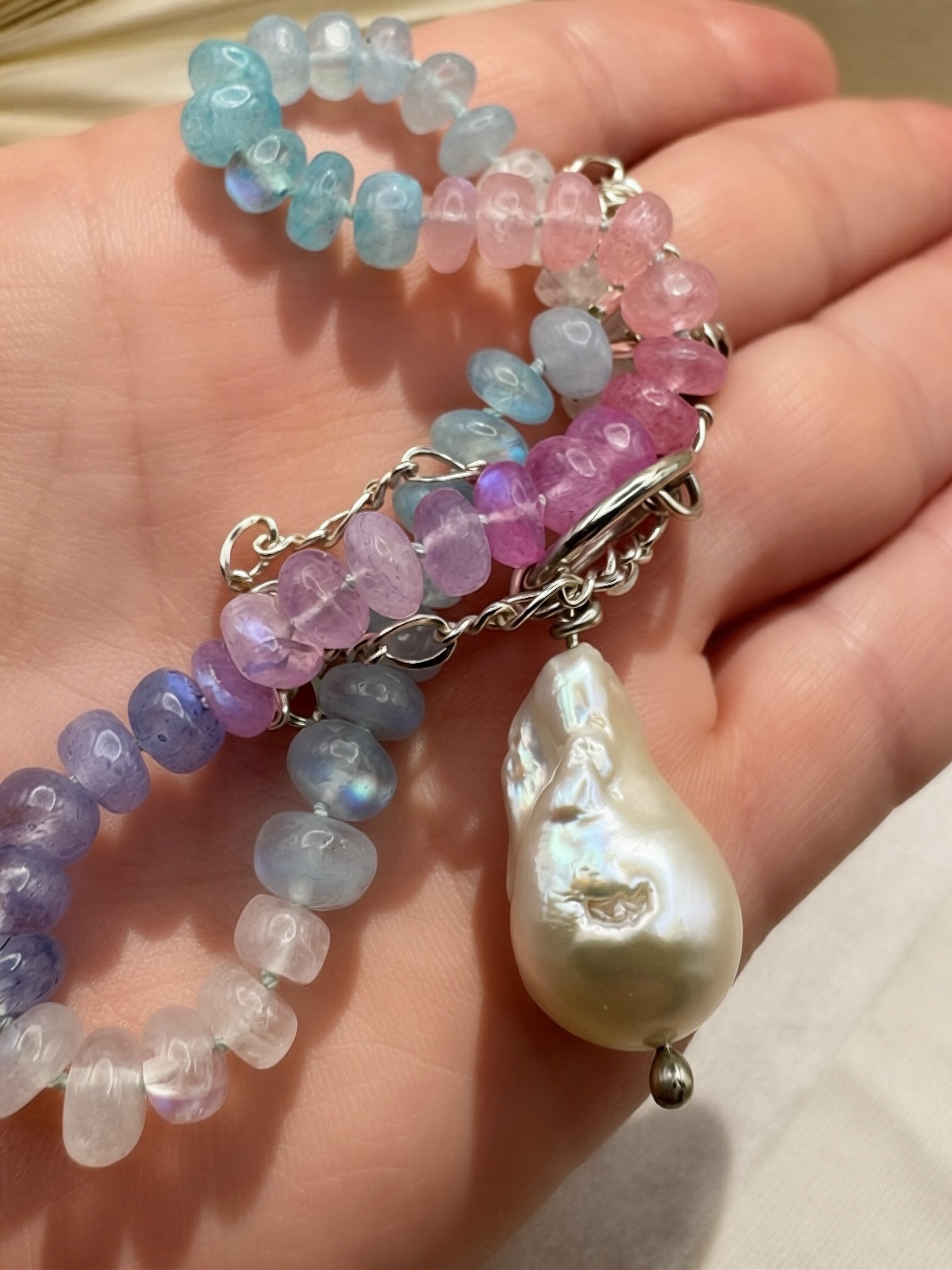 A close-up of a beaded necklace draped across a hand. The bracelet features translucent moonstone round beads in pastel shades of blue, pink, purple, and white, connected by a silver chain with a large, lustrous teardrop-shaped baroque pearl pendant.