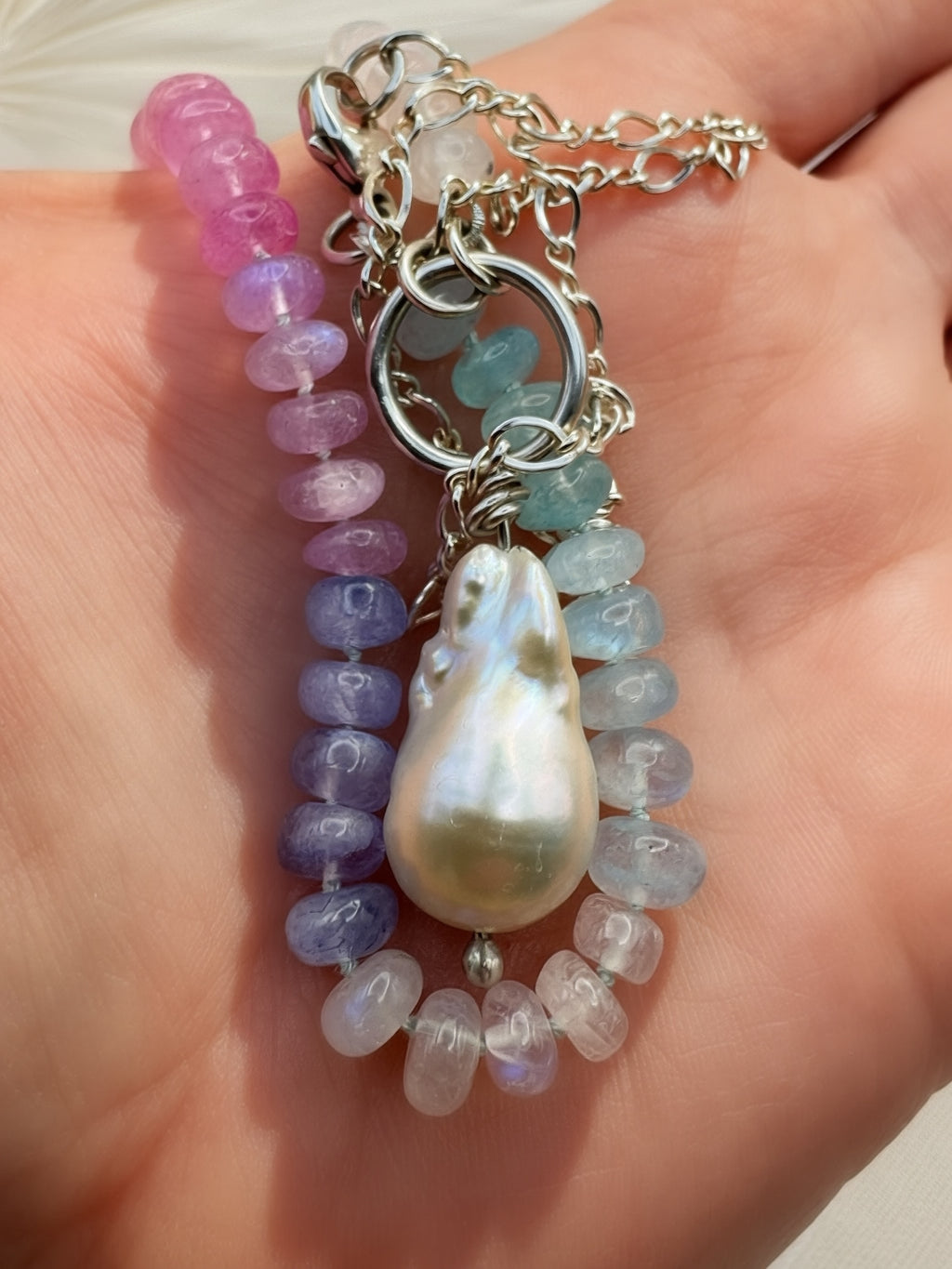 A close-up of a hand holding a beaded and silver necklace which features a large baroque pearl pendant, and strands of translucent moonstone beads in pink, purple, blue, and white, with a silver chain.