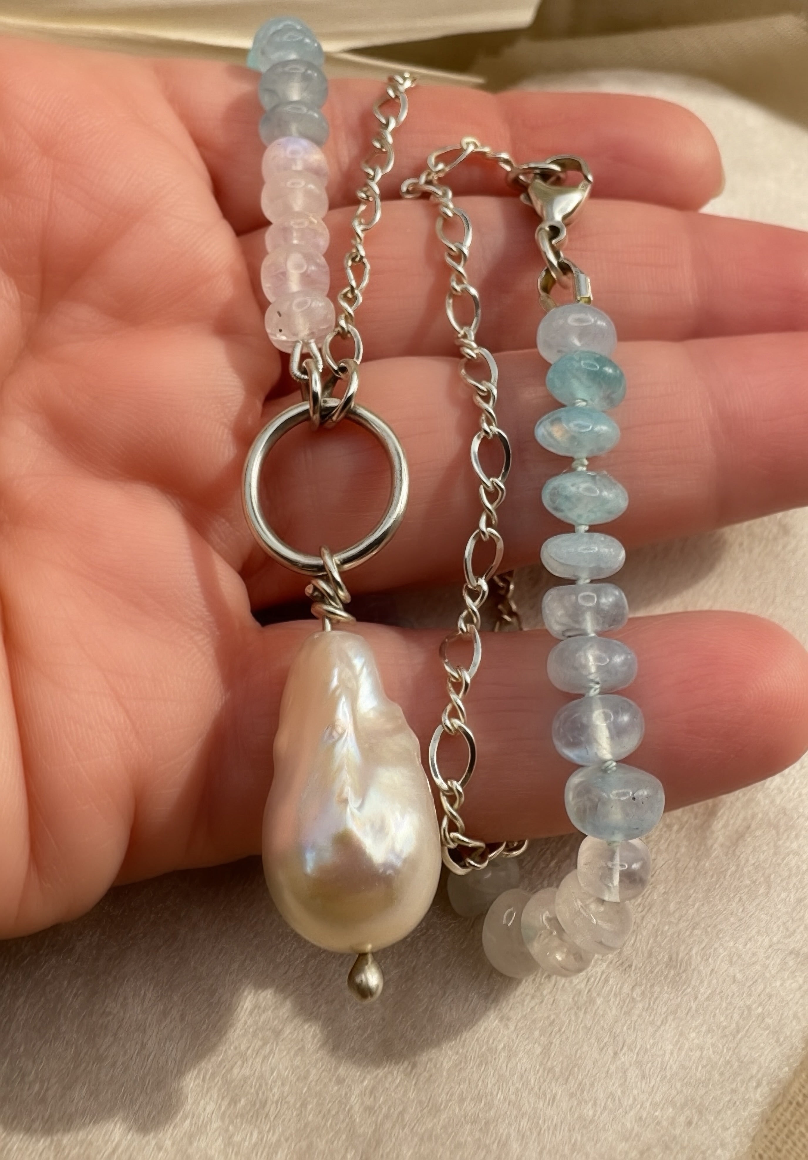 A close-up of a hand holding a silver chain necklace featuring a large baroque pearl, a round silver ring, and beaded strands of light blue, pink, and clear faceted moonstone beads.