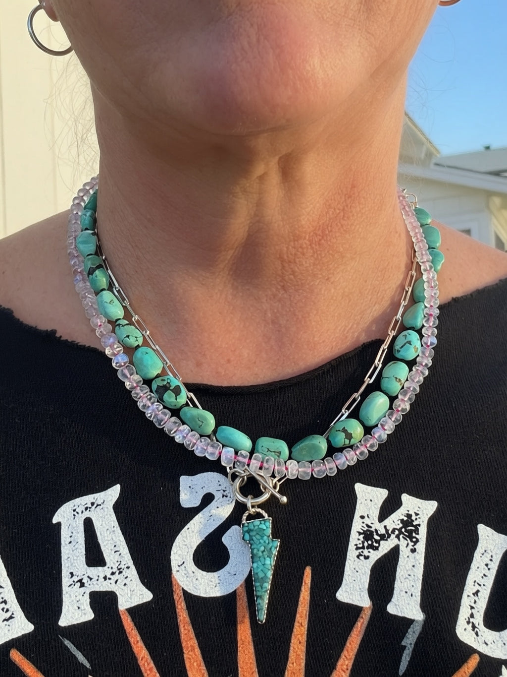 A close-up of a person wearing layered necklaces with turquoise beads, clear faceted moonstone beads, and a dangling turquoise lightning bolt charm, paired with a black graphic shirt.