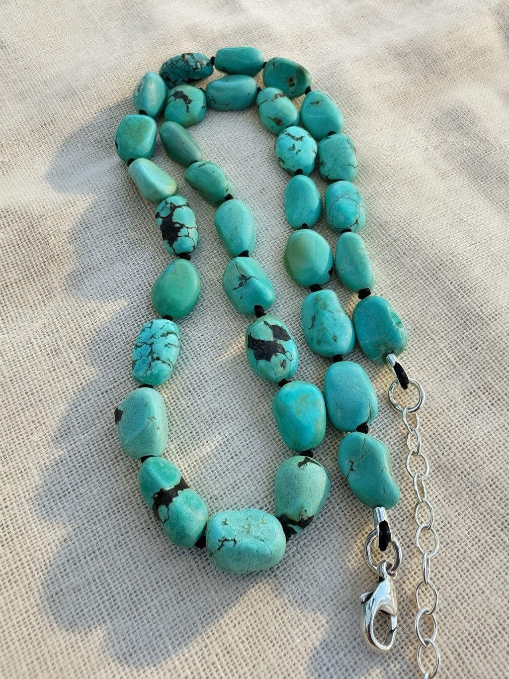 A beaded necklace made of turquoise beads with black matrix, laid on a beige fabric background. The necklace has a silver chain extender and lobster clasp.
