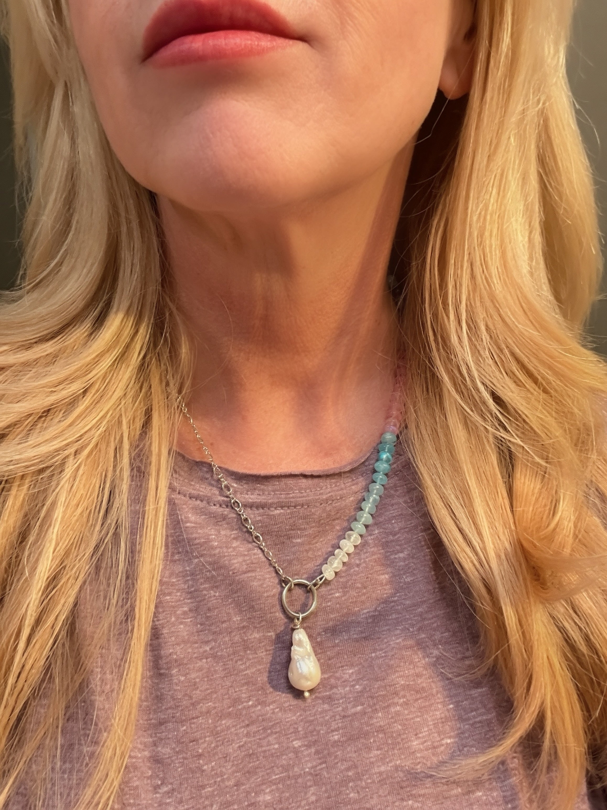 A close-up of a woman wearing a half sterling silver chain, half beaded necklace with a baroque teardrop-shaped pearl pendant, and a beaded chain featuring pastel rainbow beads