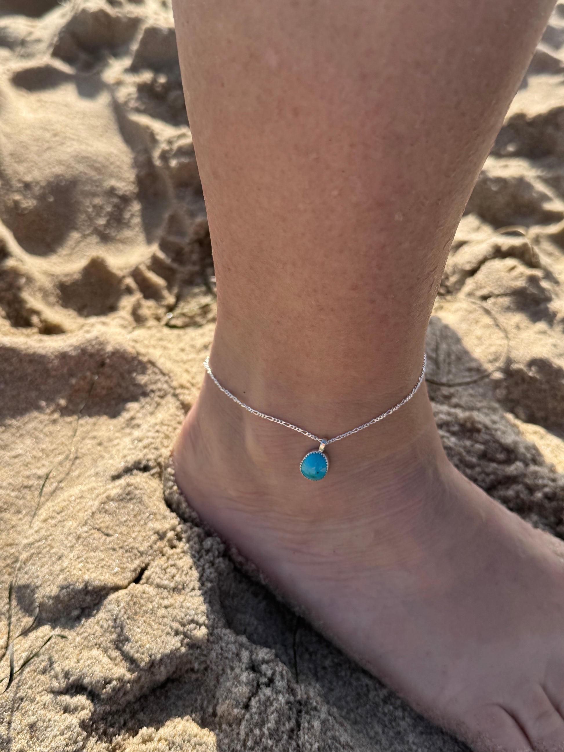 Tear drop shaped Bisbee turquoise anklet on a silver Figaro chain On an ankle against a sandy background
