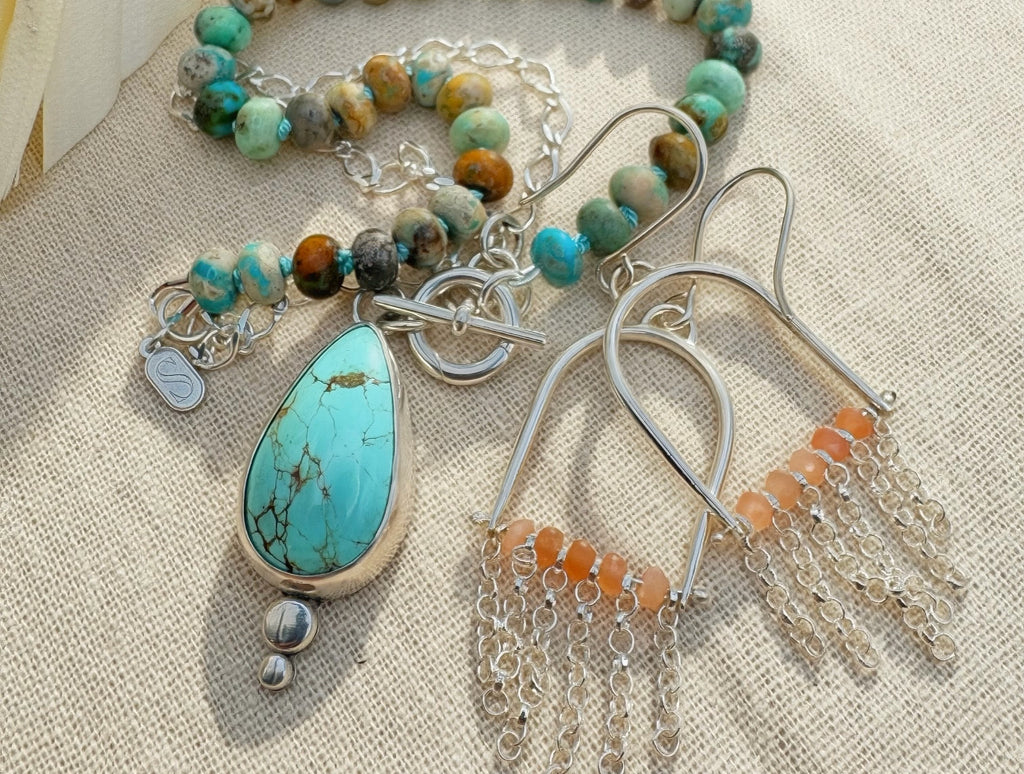 A jewelry set laid flat on a beige fabric background, featuring a turquoise teardrop pendant necklace, silver arch earrings with peach moonstone beads and sterling fringe.