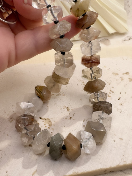 Close-up of faceted rutilated quartz beads in clear, smoky, champagne and earthy tones, hand-knotted on deep graphite silk.